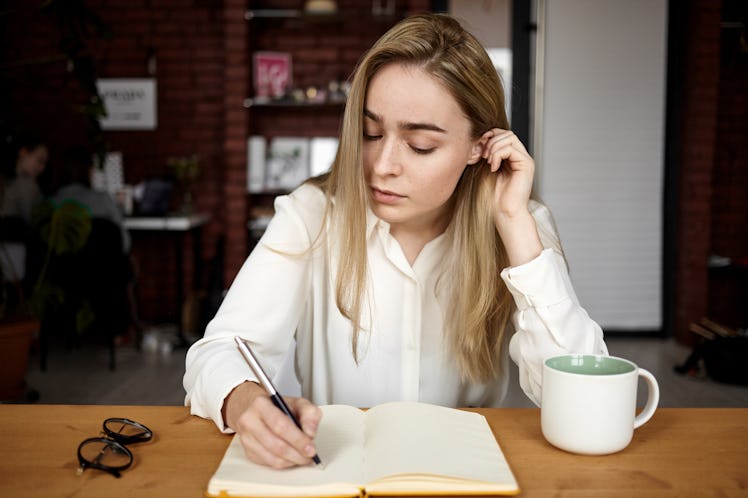 Candid shot of attractive blonde student girl in white blouse doing homework at workplace at home, w...