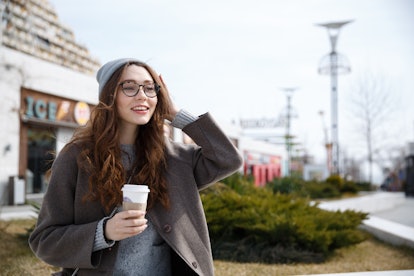 Smiling cute young woman walking and drinking take away coffee outdoors