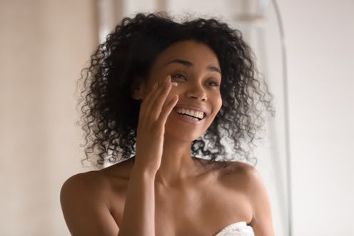 Smiling african American young woman wrapped in towel after shower look in mirror apply under eye an...