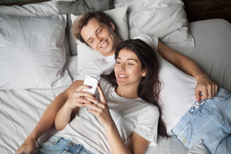 Smiling attractive millennial couple using smartphone lying on bed together, happy boyfriend and gir...