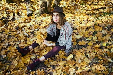 A brunette woman in maroon boots and leggings sits in a pile of yellow leaves in the fall.