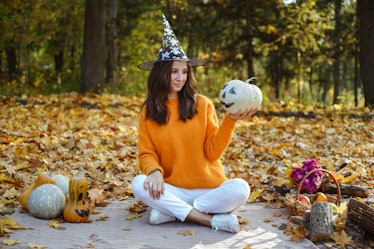 A girl sits outside on Halloween in white jeans, an orange sweater, and a witch hat and holds a pump...