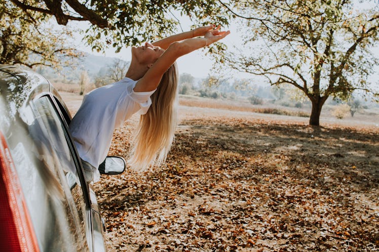 A happy blonde woman sticks her head and arms out of a parked car in the fall foliage.