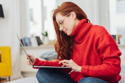 Young woman in a bright red winter sweater sitting working on a laptop on a bed at home in a close u...