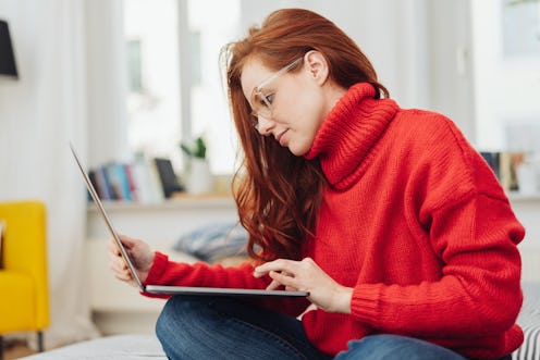 Young woman in a bright red winter sweater sitting working on a laptop on a bed at home in a close u...