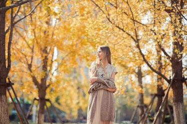 A woman with long blonde hair in a long dress and sweater walks through the yellow foliage in the fa...