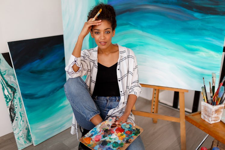 Close up portrait of charming woman with dark skin posing in her art studio with abstract paintings...
