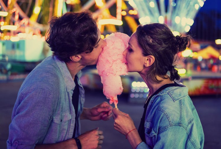 A couple having a date in a theme park like Disneyland share pink cotton candy in front of a Ferris ...