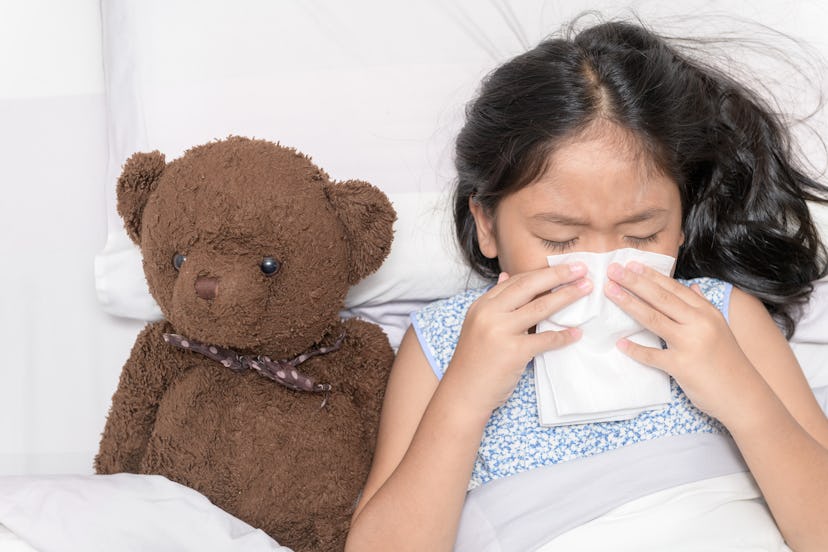 Sick little girl with the flu, blowing her nose into tissue lying with her teddy bear in her bed