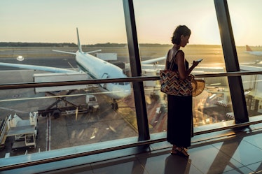 A woman with short hair and a long dress stands by the window in an airport and checks her phone whi...