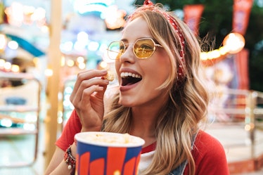 A happy blonde woman wearing sunglasses and a red T-shirt smiles while eating kettle corn in an amus...