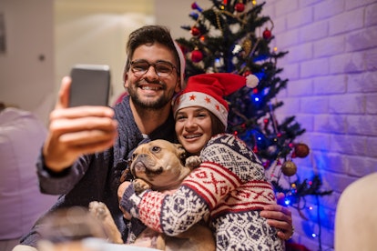 A smiling couple takes a selfie with their dog while wearing Christmas sweaters. 