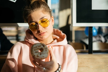 A girl in a pink sweatshirt and yellow sunglasses looks at a pumpkin doughnut in her hand while sitt...