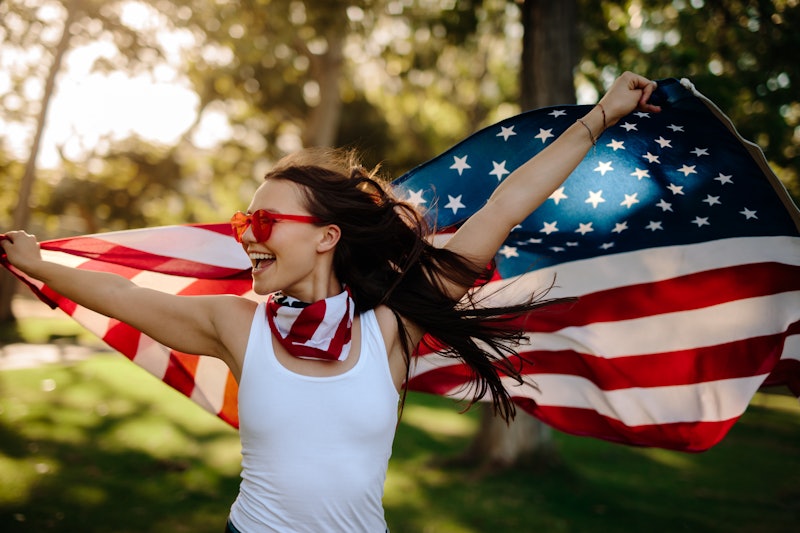 Young woman enjoying in park holding USA flag. American girl with national flag having fun at the pa...