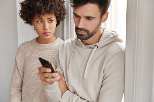 Attractive man wears hoodie, ignores live communication with wife, focused in modern smart phone, re...