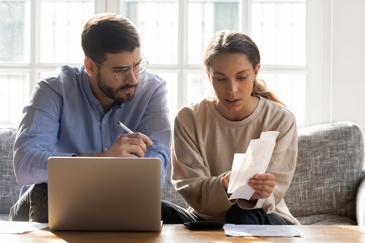 Serious couple calculating expenses, bills together, using laptop, sitting on couch at home, checkin...