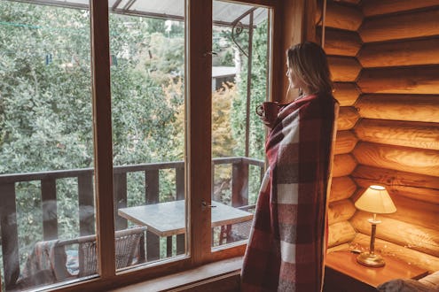 Woman in warm blanket relaxing and drinking morning coffee on cozy bed in log cabin in winter