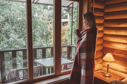Woman in warm blanket relaxing and drinking morning coffee on cozy bed in log cabin in winter