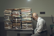 Desperate depressed businessman with lots of paperwork in his messy office at night, he is leaning o...