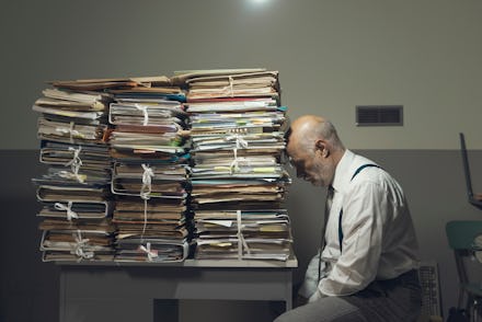 Desperate depressed businessman with lots of paperwork in his messy office at night, he is leaning o...