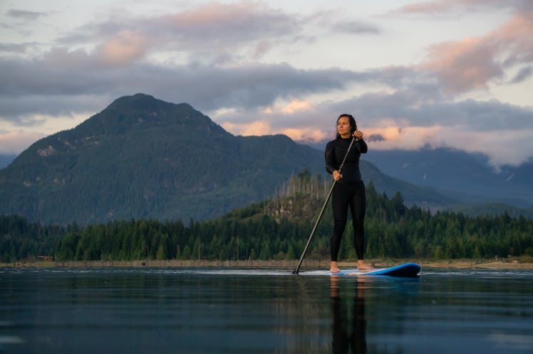 Adventurous Girl on a Paddle Board is paddling in a calm lake with mountains in the background durin...