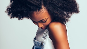 Shy little African girl with long curly hair wearing dungarees standing sideways alone against a gra...