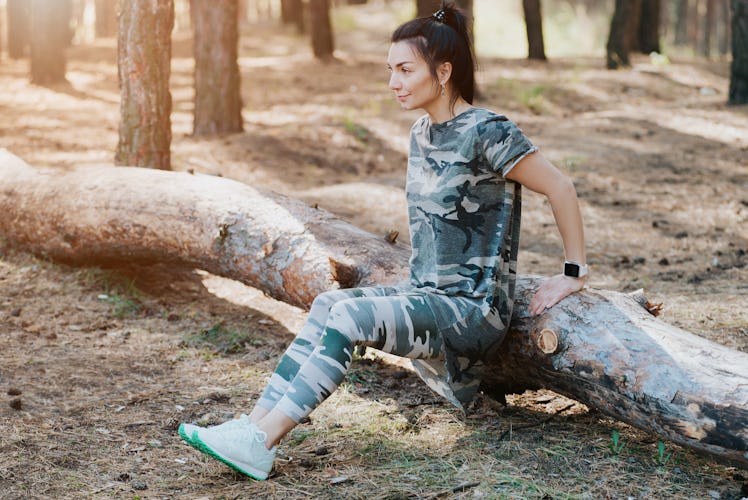 A brunette woman wearing camo shirt and leggings works out in the woods.