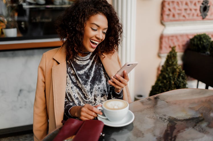 A stylish girl laughs while looking at her phone in a cute café with a pumpkin coffee on the table.