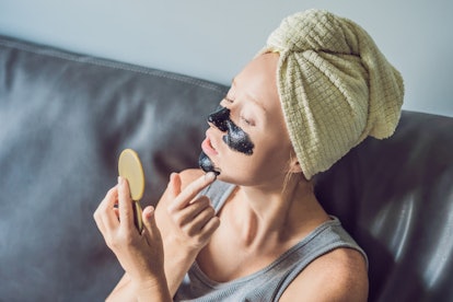 Beautiful young woman relaxing with face mask at home. Happy joyful woman applying black mask on fac...