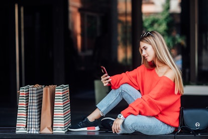 Beautifulblonde woman sitting on a street and surrounded by shopping bags and looking on phone.