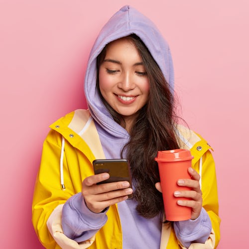 Indoor shot of happy dark haired girl wears hoody and raincoat, holds mobile phone, scrolls feed in ...