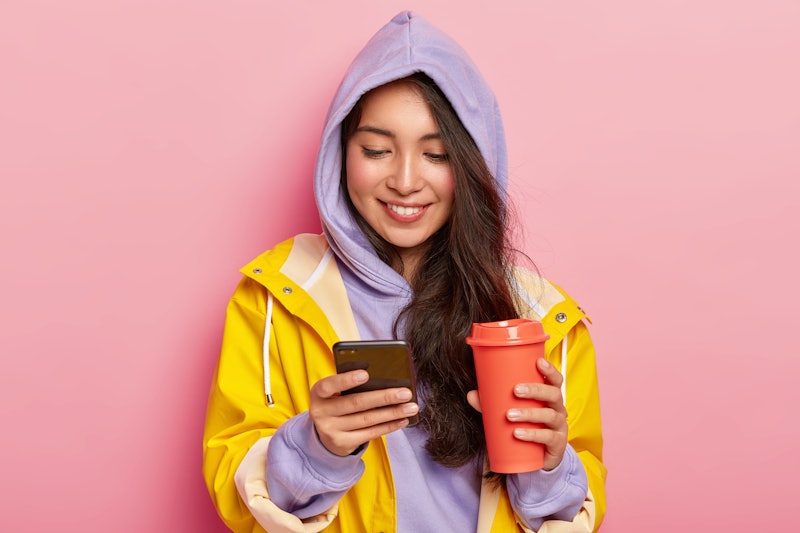 Indoor shot of happy dark haired girl wears hoody and raincoat, holds mobile phone, scrolls feed in ...