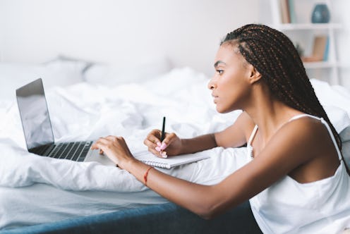concentrated african american woman with laptop in bed writing in notepad