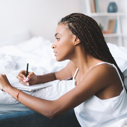 concentrated african american woman with laptop in bed writing in notepad