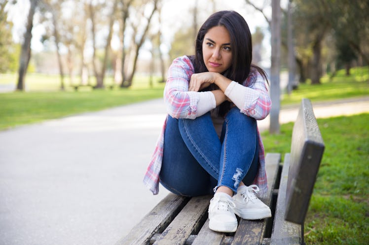 Pensive young woman contemplating in park. Serious sad mixed race girl sitting on bench and looking ...