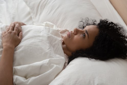 Thoughtful african American millennial girl lying in white comfortable bed lost in thoughts think of...