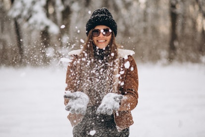 A woman in sunglasses, beanie cap, and jacket smiles and plays outside on a snow day. 