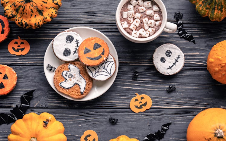 A plate of Halloween cookies on a black wooden table is next to a mug of hot cocoa topped with marsh...