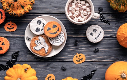 A plate of Halloween cookies on a black wooden table is next to a mug of hot cocoa topped with marsh...