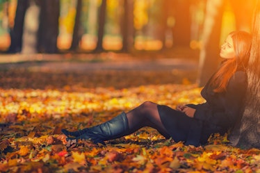 Beautiful woman in black coat and leather boots sitting under tree in autumn park with fallen leaves...
