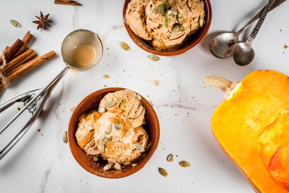 Pumpkin ice cream with pumpkin seeds on top sits in a bowl on a table next to ingredients and kitche...