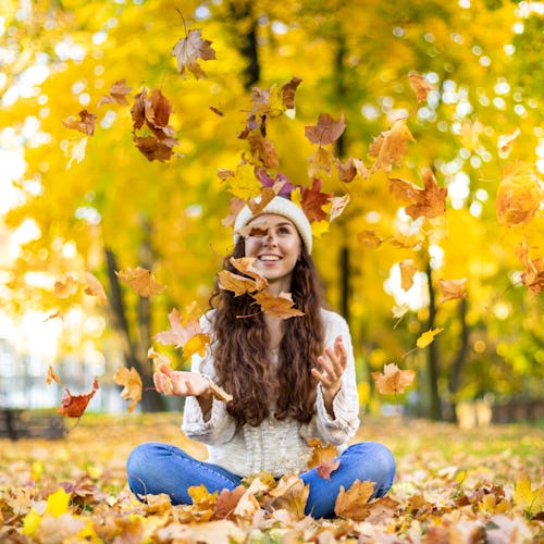 Happy young woman throwing autumn leaves and smiling on colorful nature city background