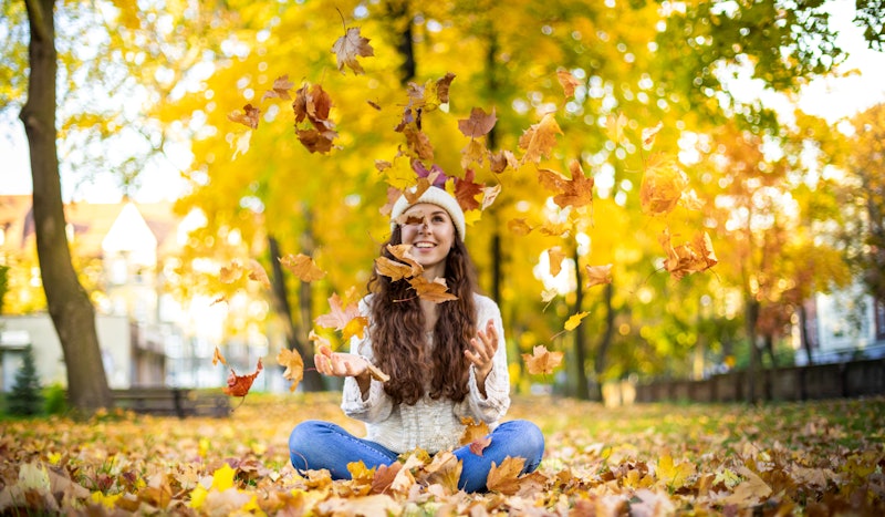 Happy young woman throwing autumn leaves and smiling on colorful nature city background