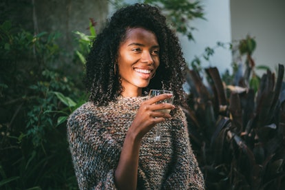 A woman in a comfy sweater smiles and holds a glass of wine outside.