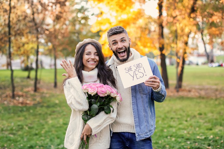 A happy couple poses for their engagement announcement showing off the ring in an autumn park.