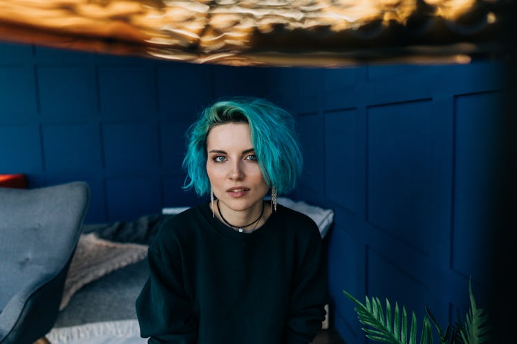 A beautiful girl in a bedroom with blue walls under a golden lamp looks directly into the frame.