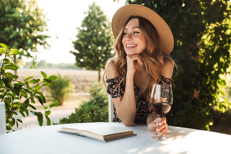 A woman on vacation sits at a table outdoors at a vineyard with a book and holding a glass of red wi...