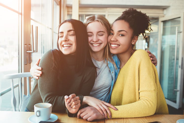 Tree young women are sitting together in a small cafe with big windows and embrace with each other.