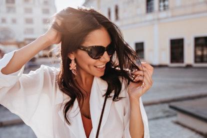 Amazing brunette woman in trendy earrings smiling on city background. Wonderful tanned girl in sungl...
