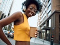 Waist up portrait of smiling woman with headphones holding coffee while walking on the street and li...